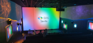 Escenario del DevFest. Pantalla gigante con el logo de GDG Sevilla sobre los colores de Google (degradado azul, rojo, verde y amarillo)